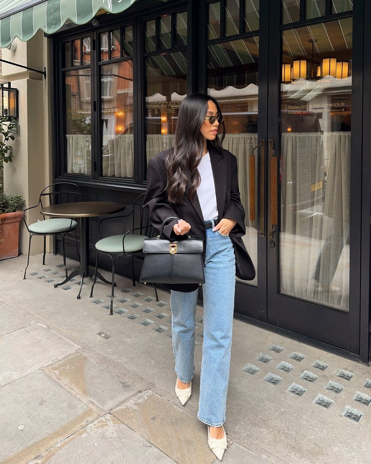 A woman wearing a black blazer, light blue jeans, and white heels walking outdoors.