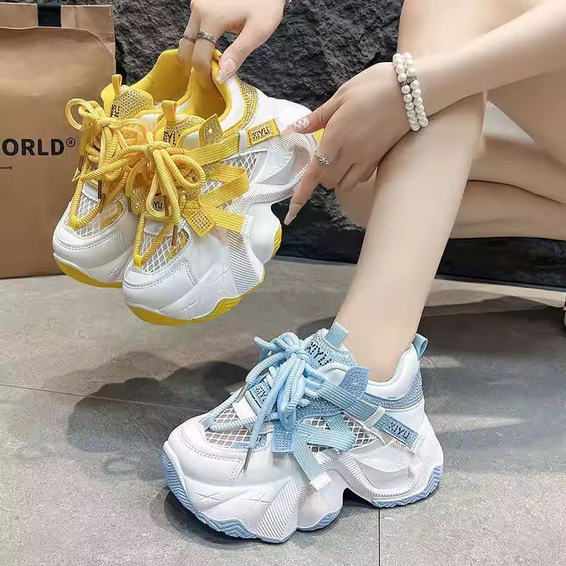 A person holding yellow chunky sneakers while wearing light blue chunky sneakers on their feet.