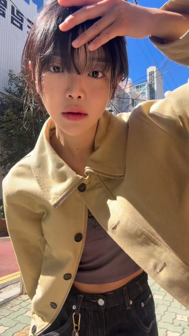 A young woman wearing a tan jacket and dark crop top posing outdoors in sunlight.