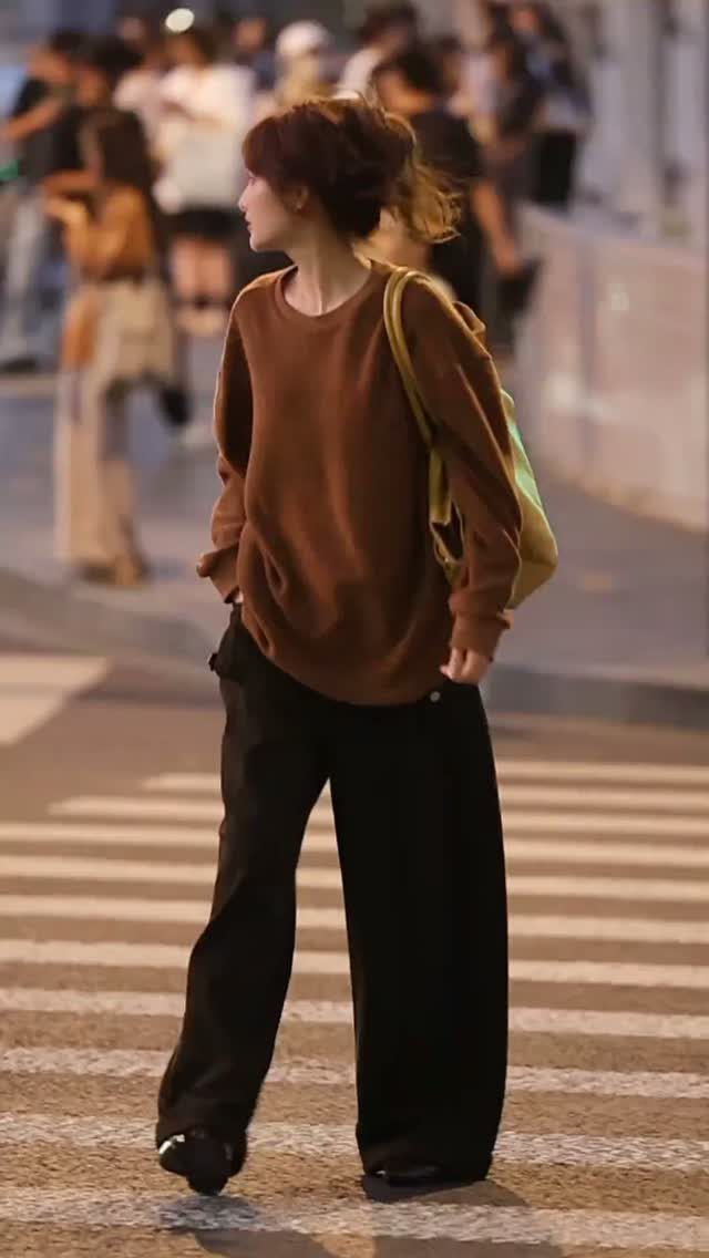 A woman in an oversized brown sweater and wide black trousers walking across a city street.