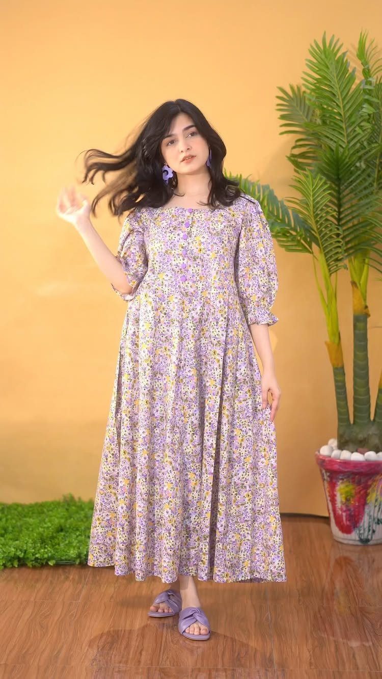 A woman wearing a long purple floral print dress standing next to a palm plant.