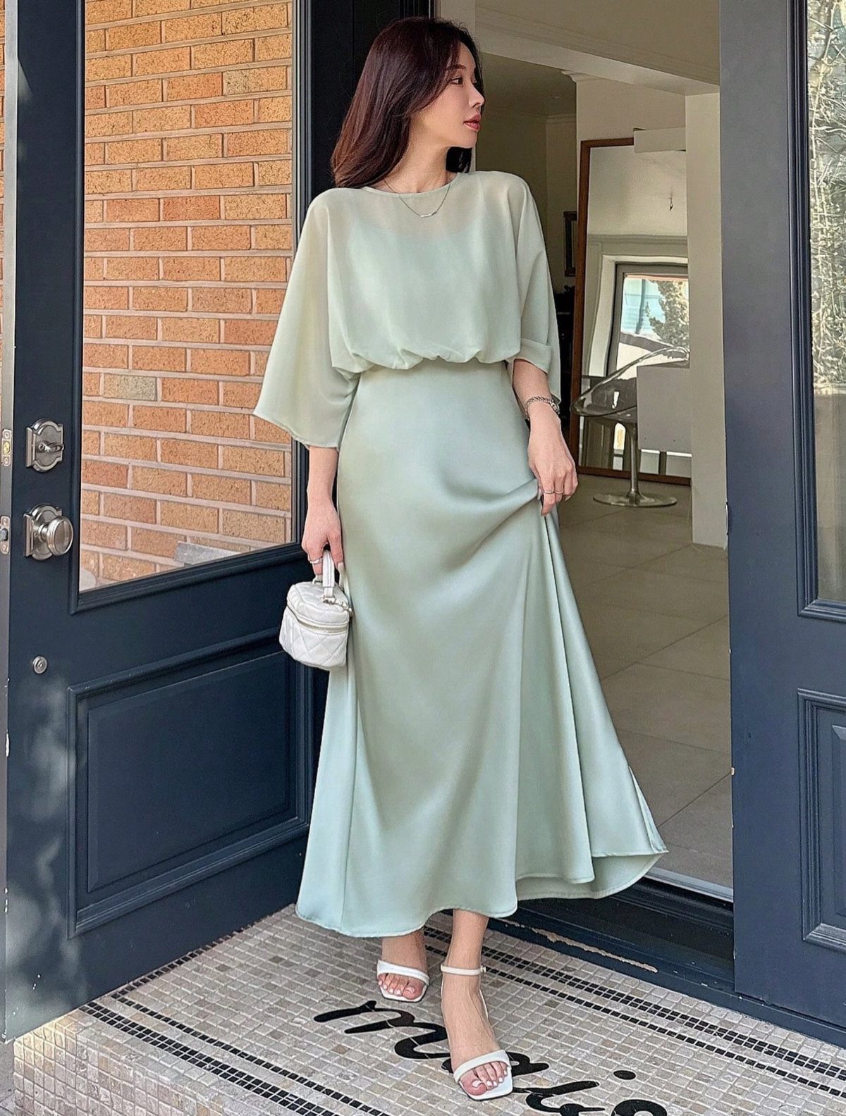 A woman in a flowing sage green satin midi dress standing in a doorway holding a white purse.