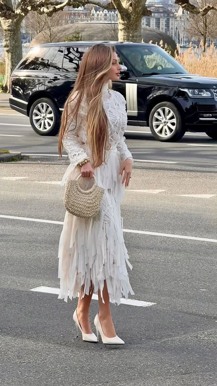 A woman in a white fringed midi dress and white heels standing on a city street.