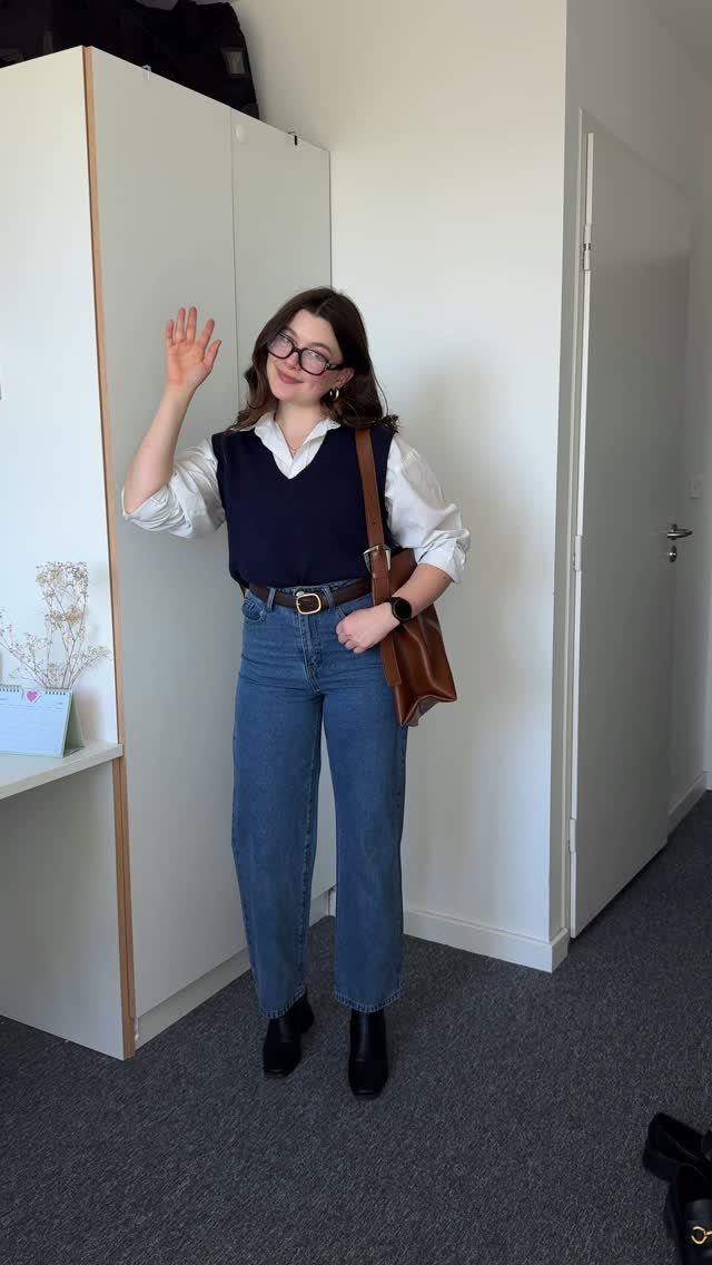 A woman in a white blouse, navy sweater vest, and blue jeans waving.