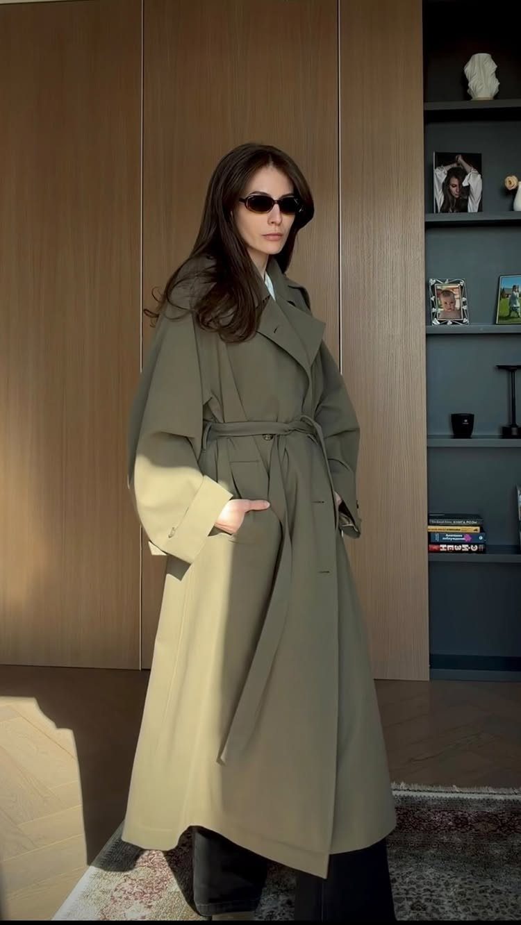 A woman wearing an olive green belted trench coat and sunglasses standing indoors.