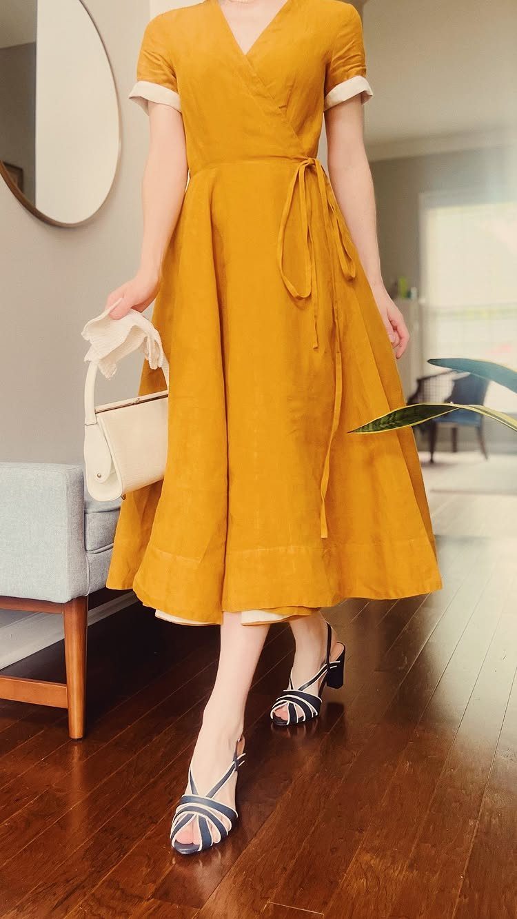 A person wearing a mustard yellow linen wrap dress and blue sandals standing indoors.