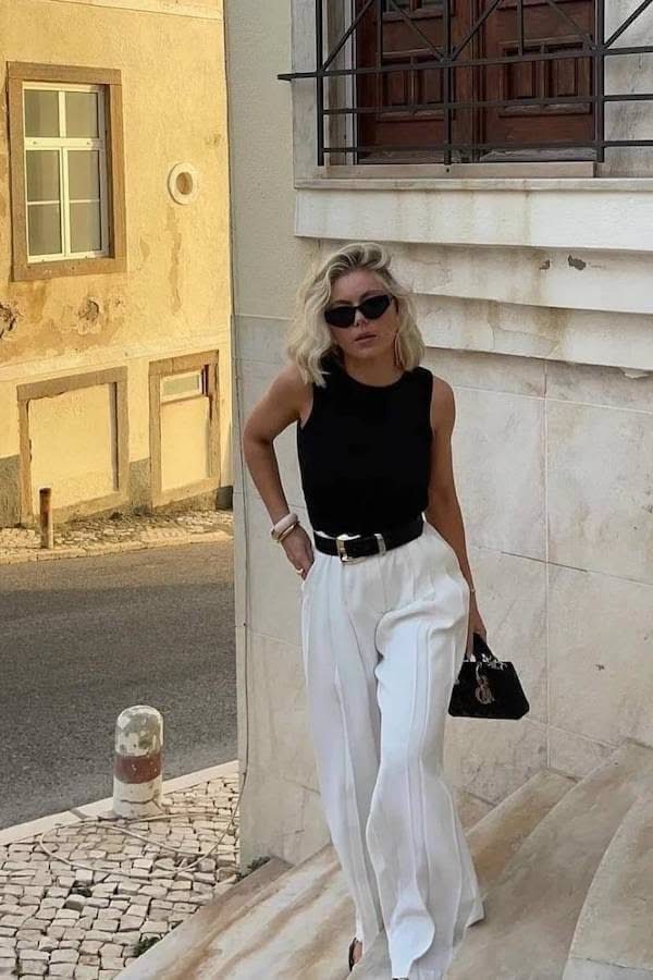 Woman wearing a black sleeveless top and white wide-leg trousers standing by a stone wall.