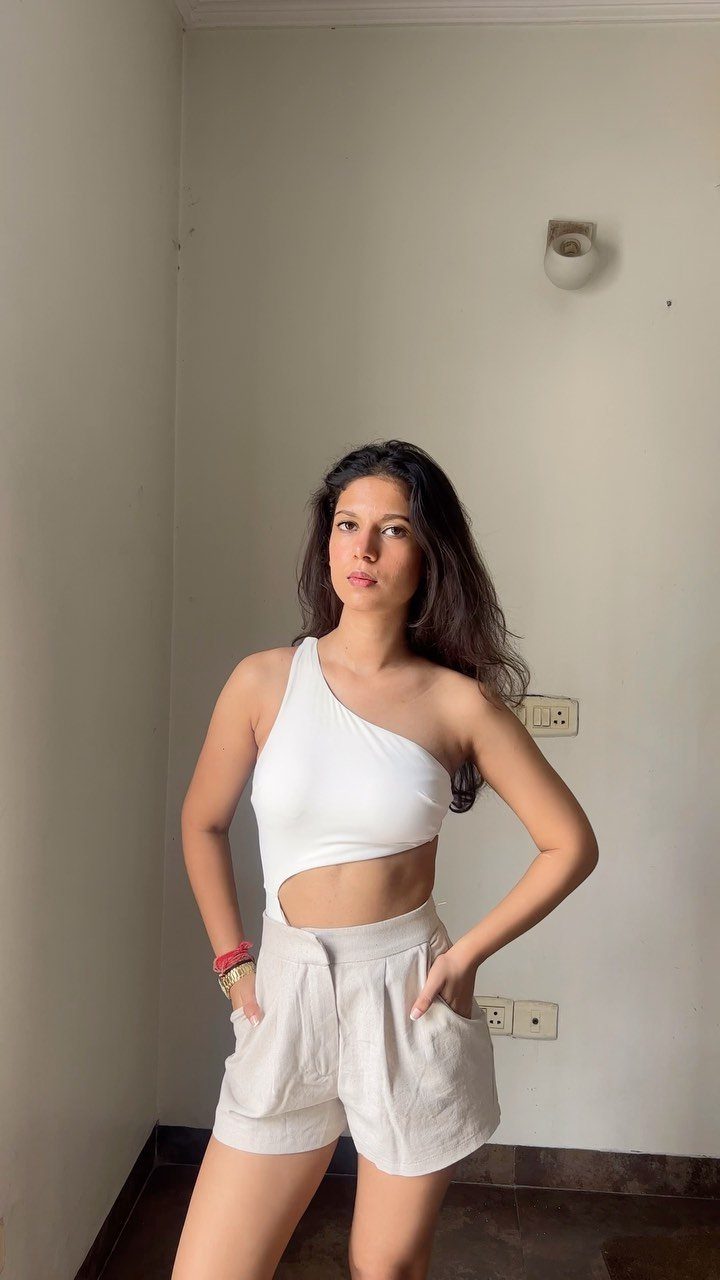 A woman wearing a white one-shoulder top and beige linen shorts standing against a white wall.