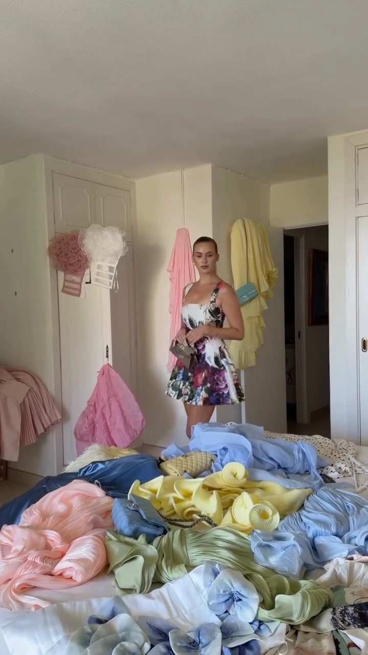 A woman standing in a room amidst piles of colorful clothing on a bed.