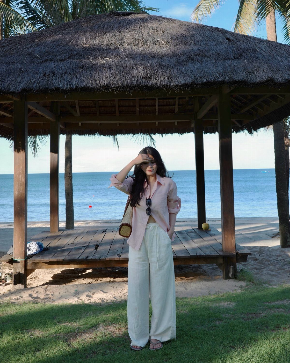 A woman wearing wide-leg white linen pants and a pink shirt standing near a beach gazebo.