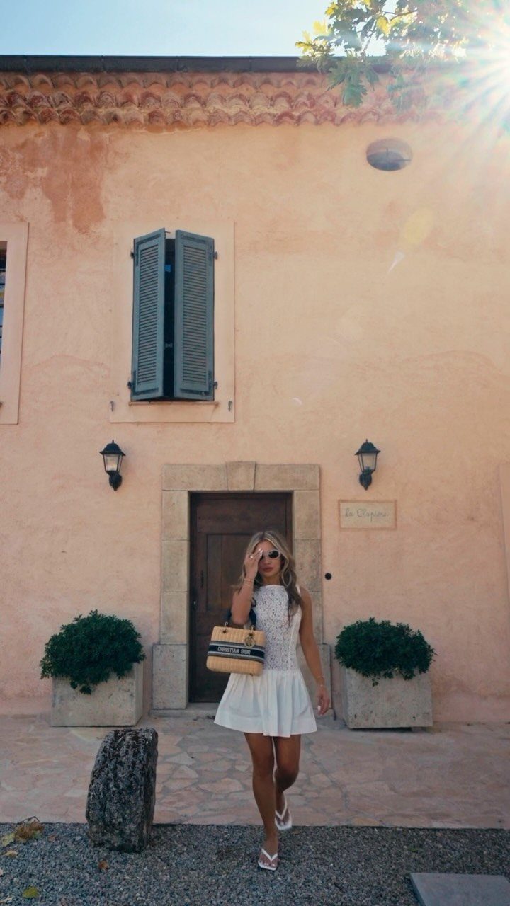 Woman in a white sleeveless mini dress and sunglasses standing before a peach building.