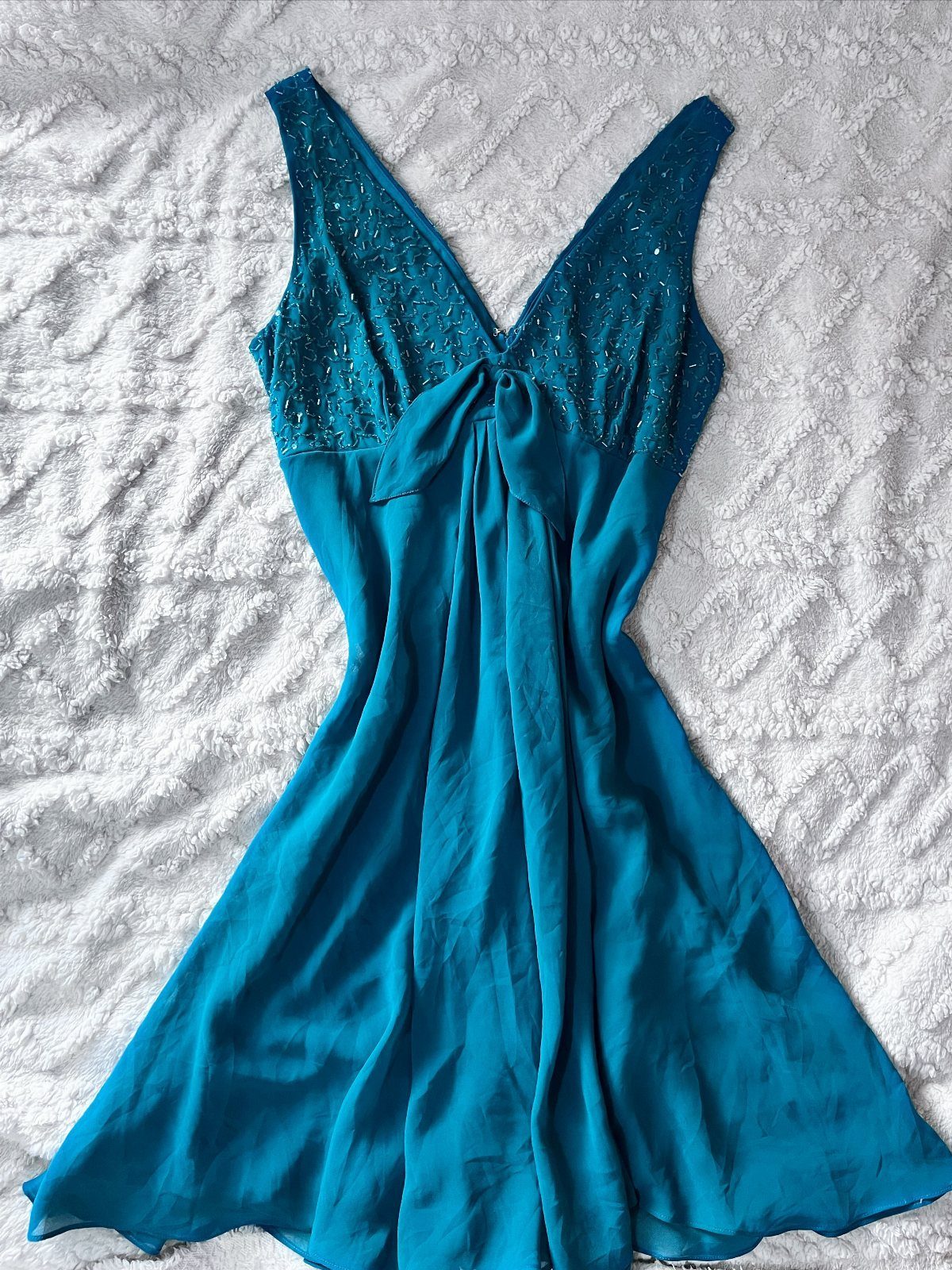 Teal sleeveless dress with beaded bodice lying on a white textured fabric background.