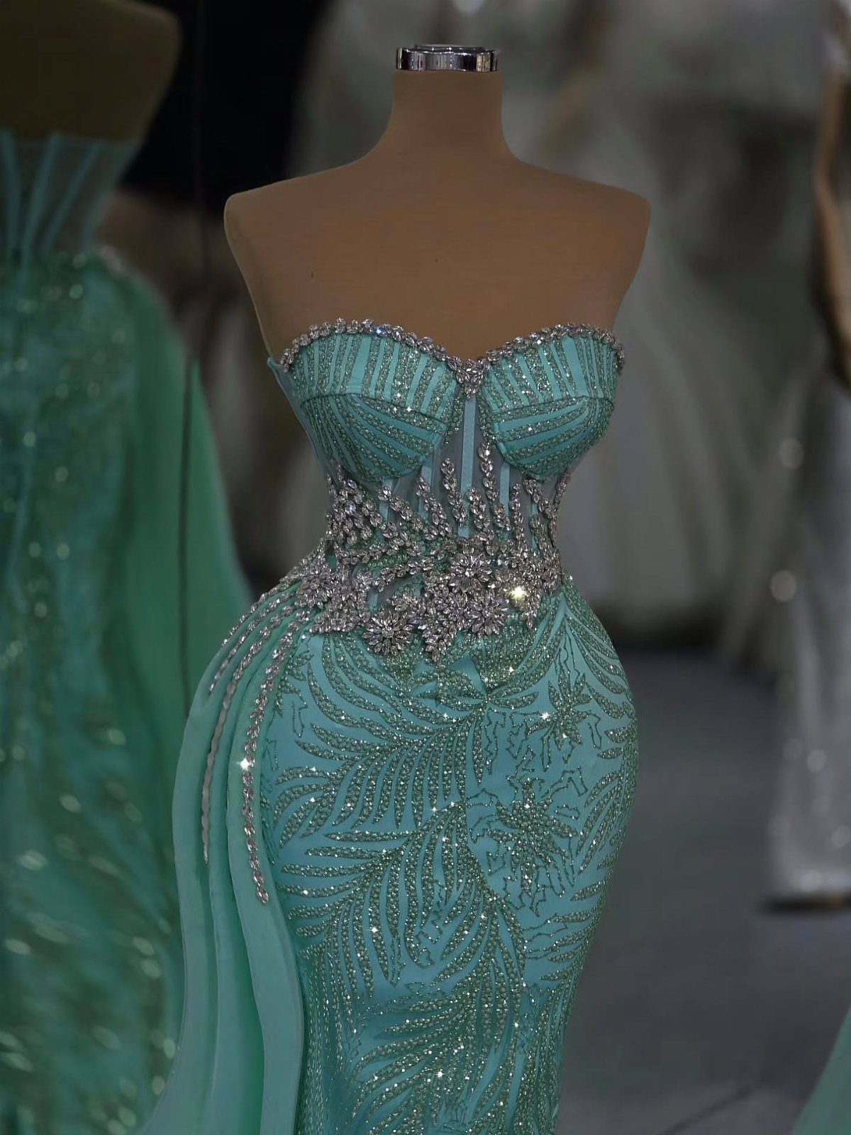 A teal strapless gown with intricate silver beadwork and crystal embellishments displayed on a mannequin.