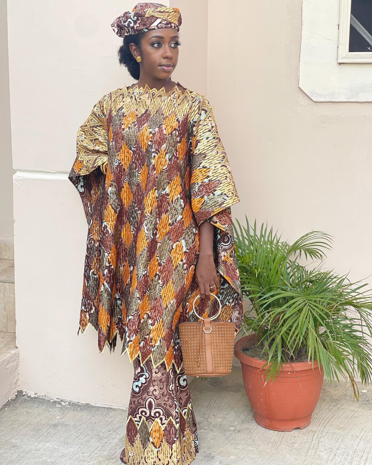 A woman wearing a patterned gold and orange Grand boubou and matching headwrap standing outdoors.