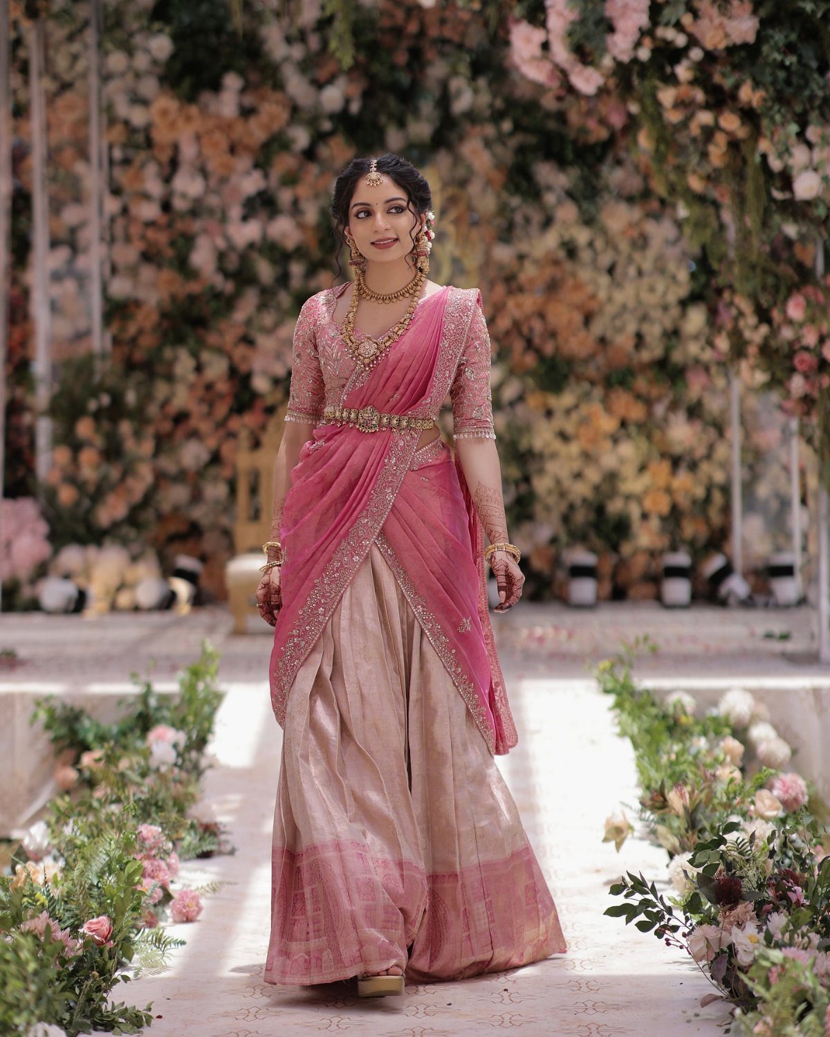 A woman wearing a pink and gold traditional South Indian half-saree standing amidst flowers.