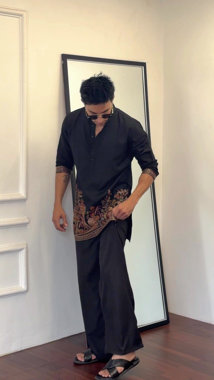 Man in black embroidered tunic and wide-leg trousers standing before a large mirror.