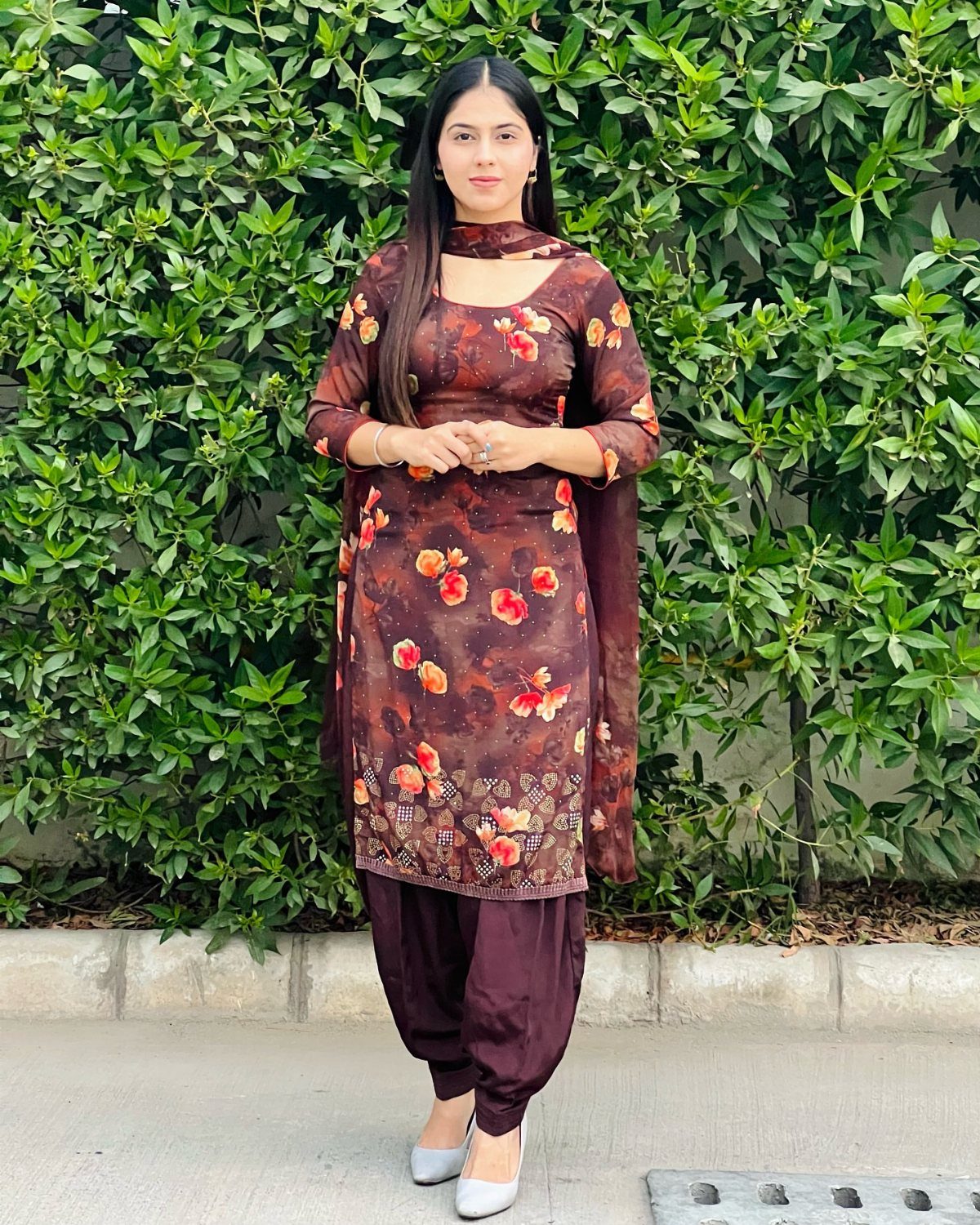 A woman posing in a floral patterned brown Salwar Kameez against a green leafy hedge.