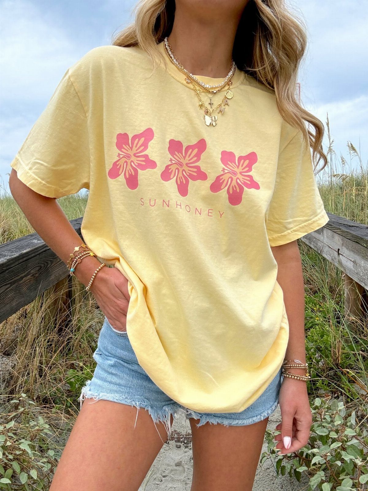 Woman wearing a yellow graphic t-shirt with pink floral designs and denim shorts outdoors.