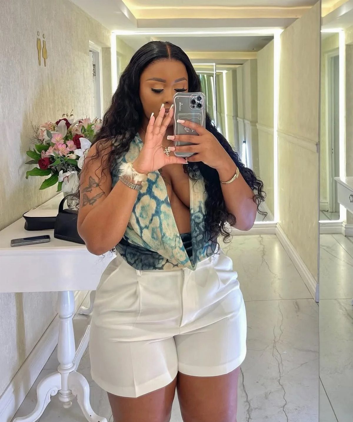 A woman posing in a mirror wearing a patterned top and white high-waisted shorts.