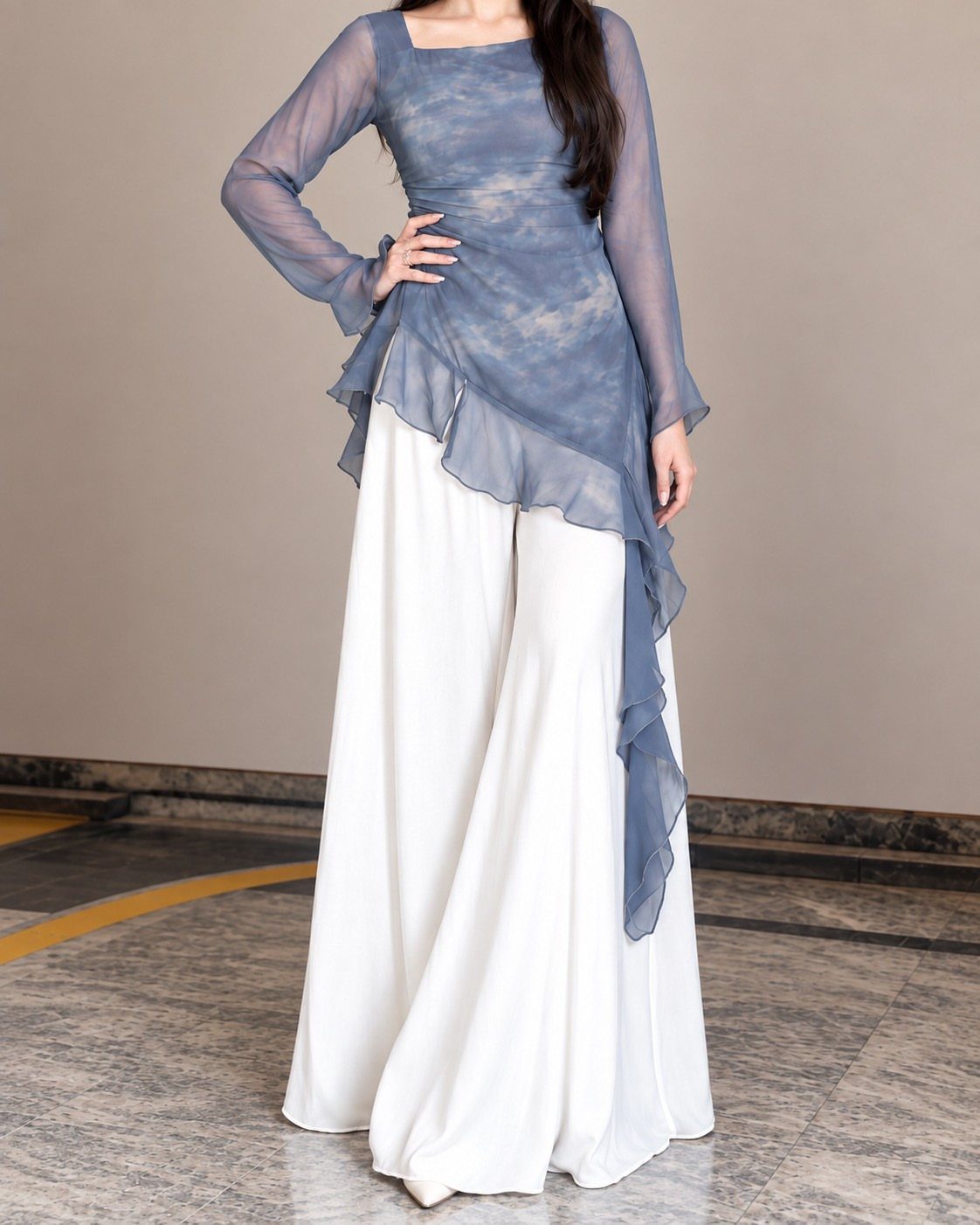 Woman wearing a blue sheer asymmetrical top and wide-leg white trousers standing indoors.