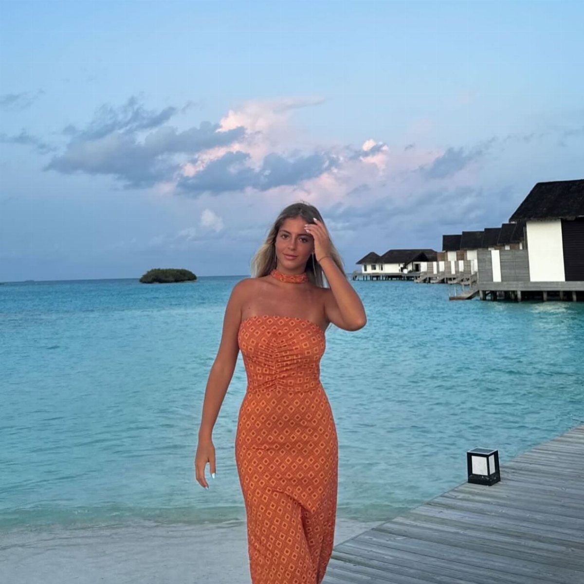 A woman in an orange patterned maxi dress standing on a pier by the ocean.