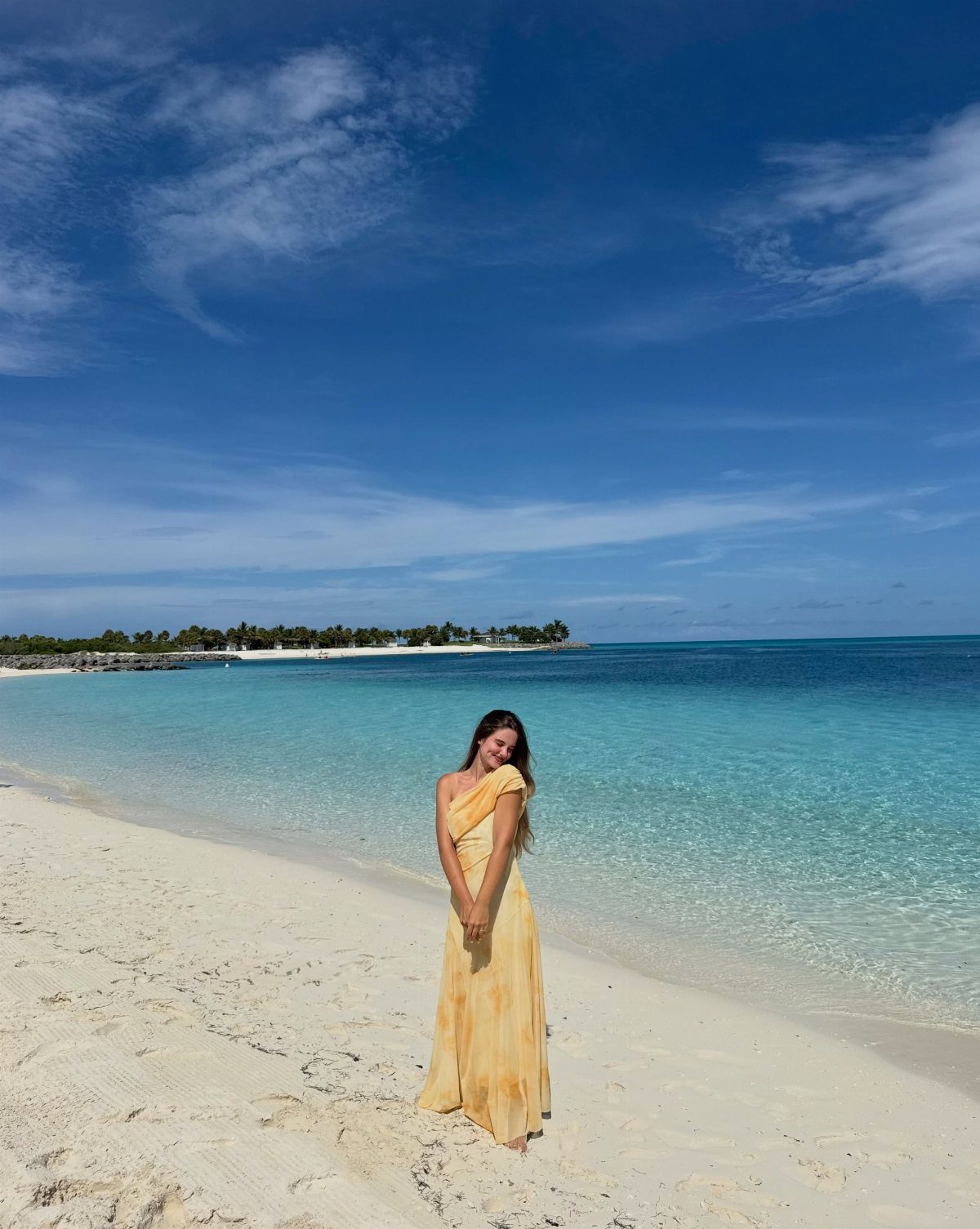 A woman in a long yellow sundress standing on a tropical beach with turquoise water.