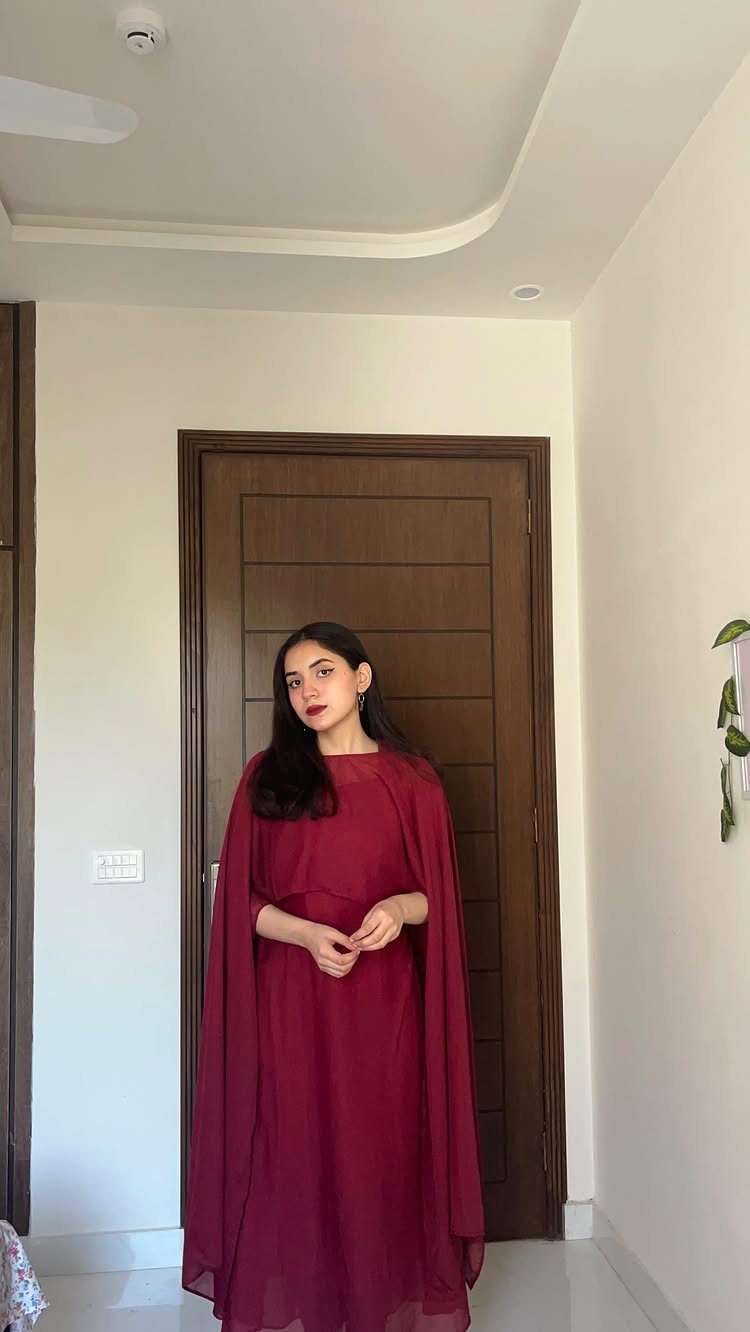 Woman in a maroon dress featuring long, flowing cape sleeves standing before a wooden door.