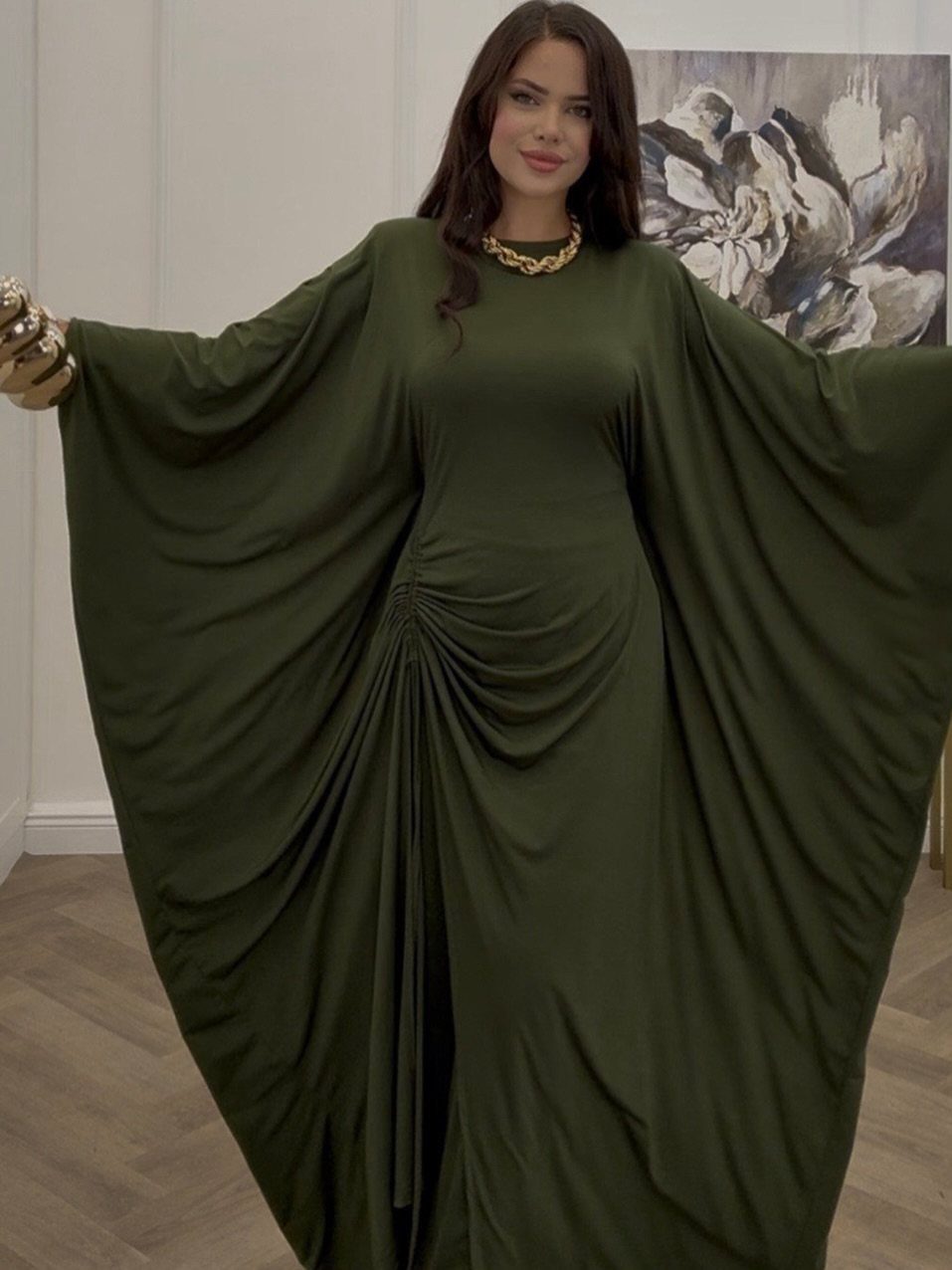 Woman in a long olive green dress with voluminous dolman sleeves and a gold necklace.