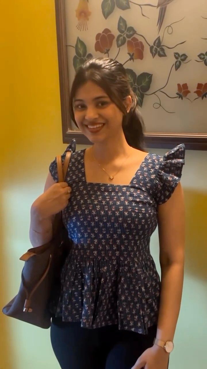 A smiling woman wearing a blue patterned top with layered frill sleeves and a brown handbag.