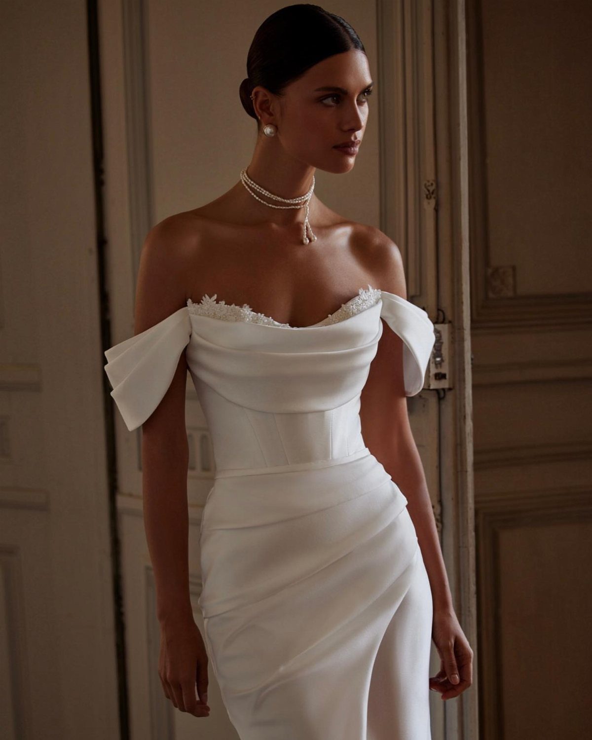 A woman in a white satin off-the-shoulder gown posing in an elegant room.