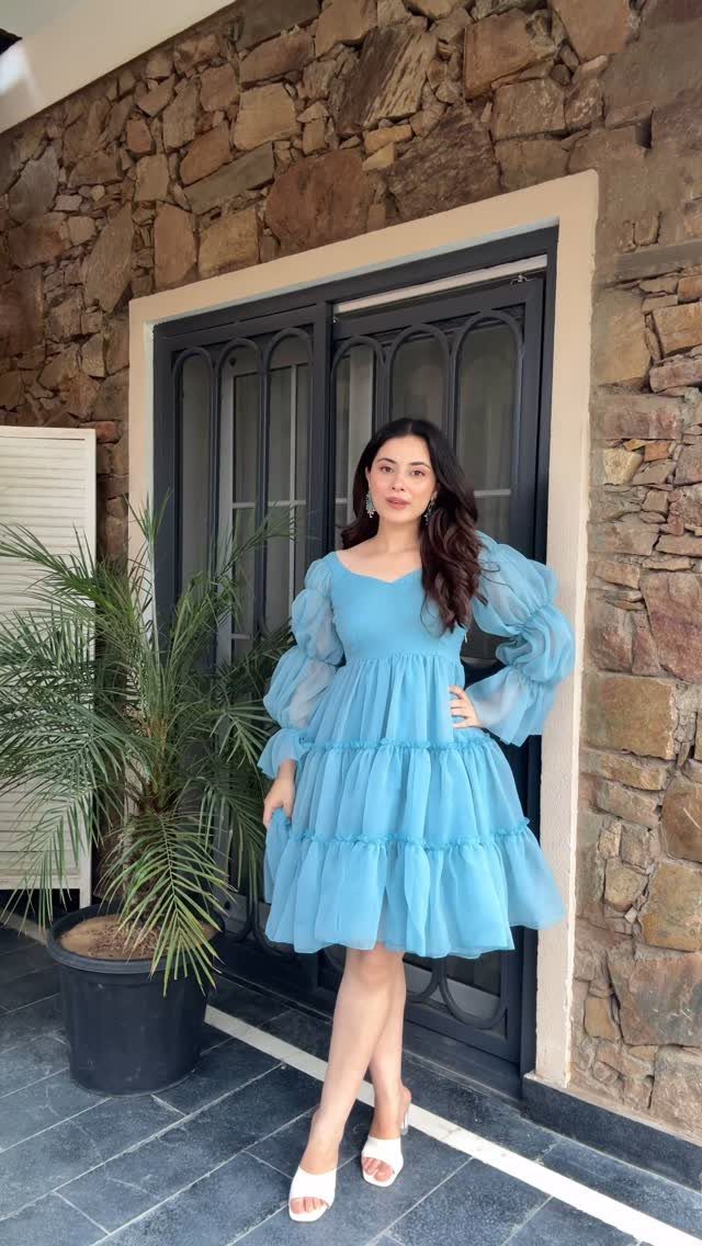 Woman in a blue tiered dress with large, sheer puff sleeves standing by a stone wall.