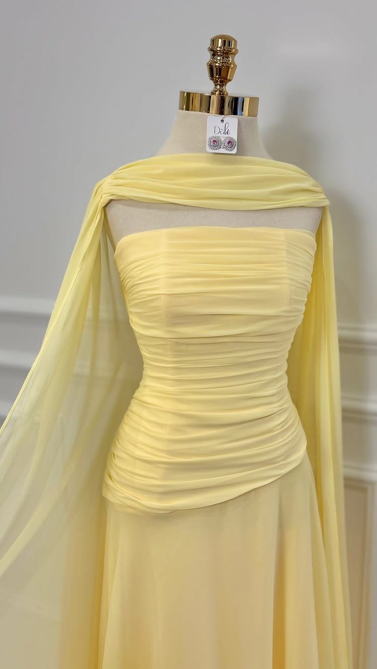 A pale yellow strapless gown with a ruched bodice and flowing chiffon cape on a mannequin.
