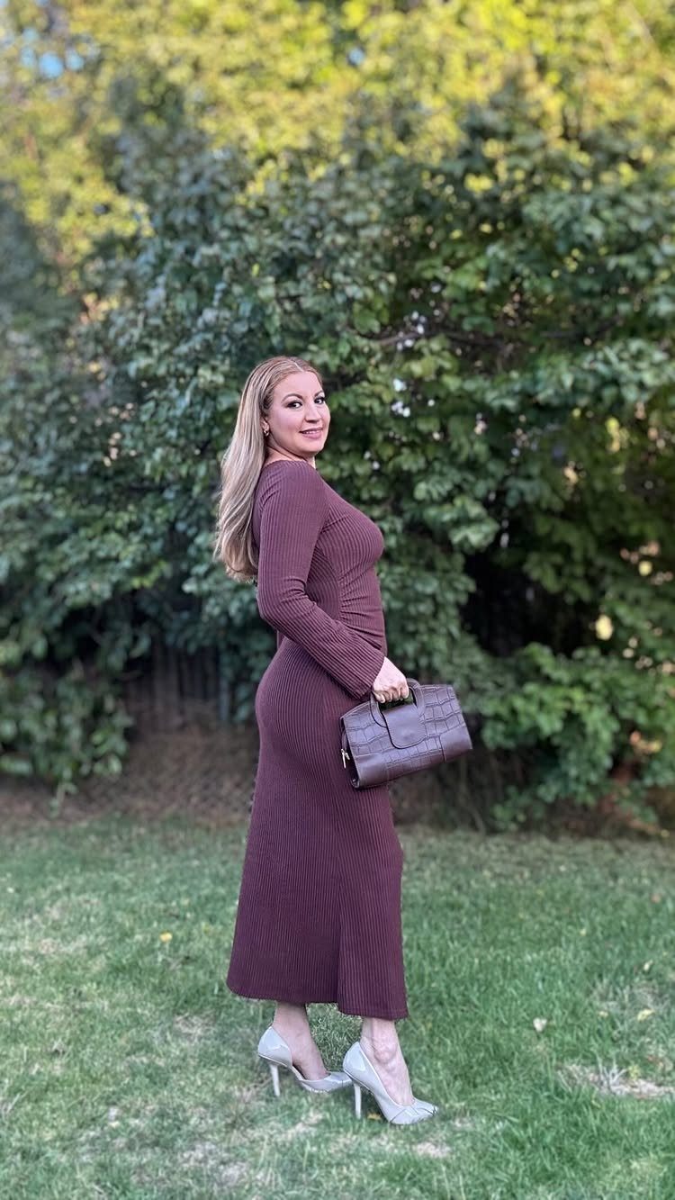 A woman posing in a long-sleeved, ribbed, burgundy bodycon midi dress and white heels.
