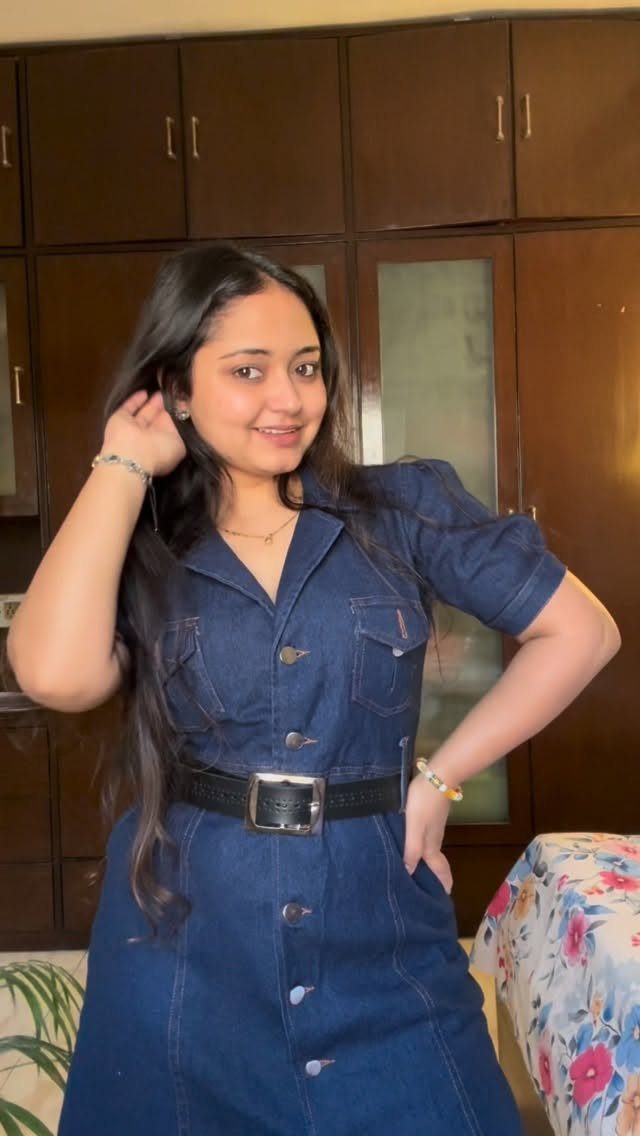 A smiling woman posing in a short-sleeved blue denim dress with a black belt.