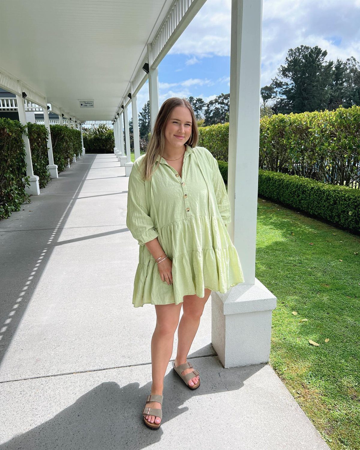 A woman smiling while wearing a light green tiered smock dress on a white walkway.