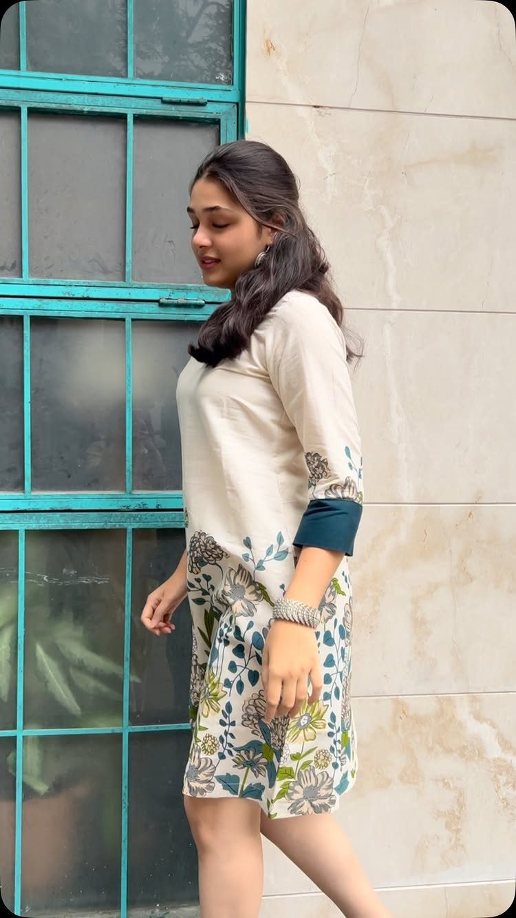 A woman wearing a cream and floral patterned tunic dress standing near a teal window.