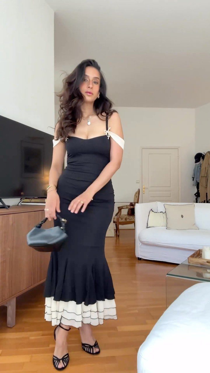 A woman posing in a black bodycon maxi dress with white ruffled trim and black sandals.
