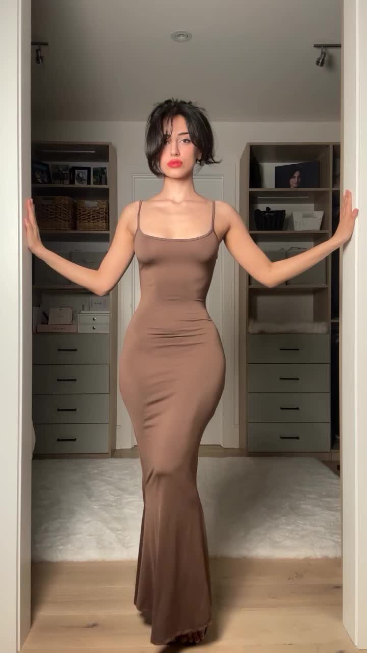 A woman posing in a brown, form-fitting, floor-length slip maxi dress with thin straps.