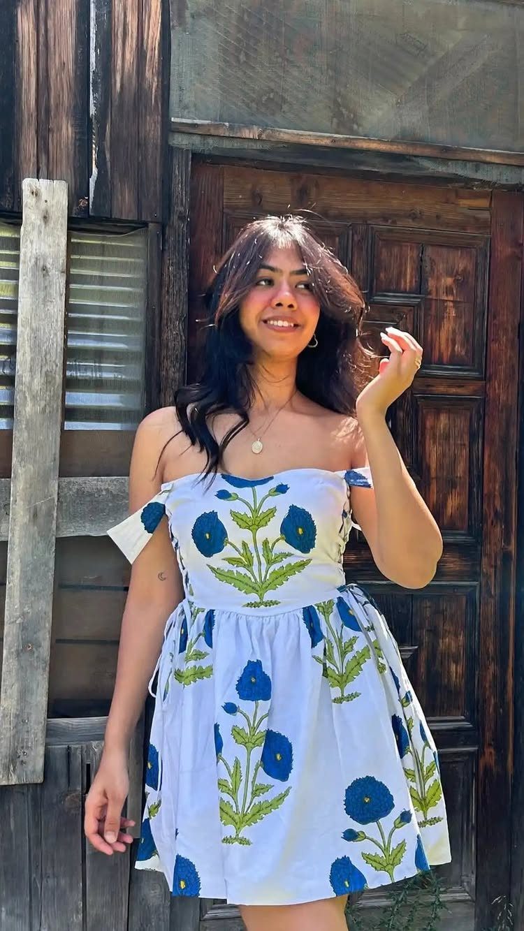 A smiling woman wearing a white off-the-shoulder dress with blue floral patterns standing outdoors.