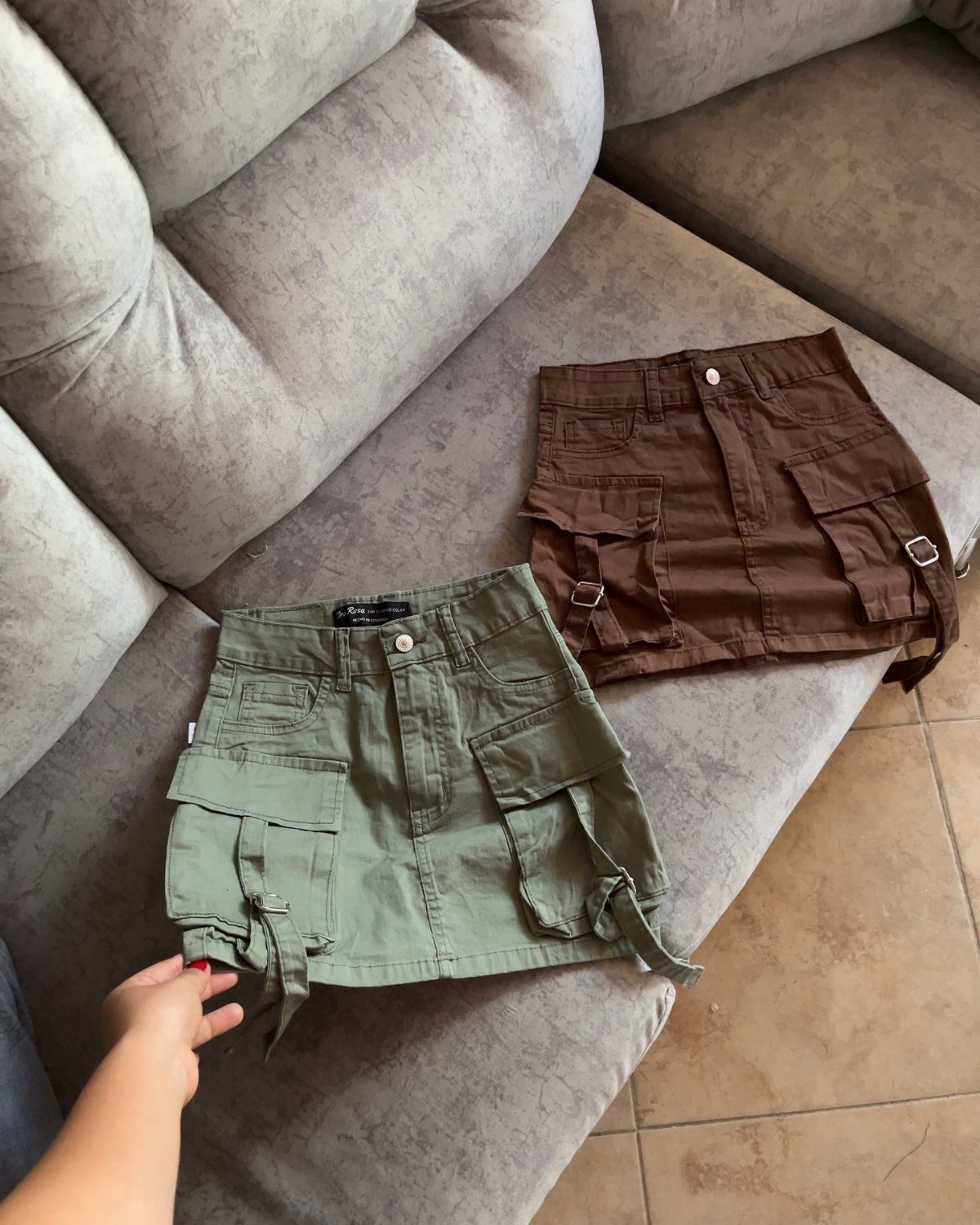 Two mini cargo skirts in sage green and brown resting on a grey tufted sofa.