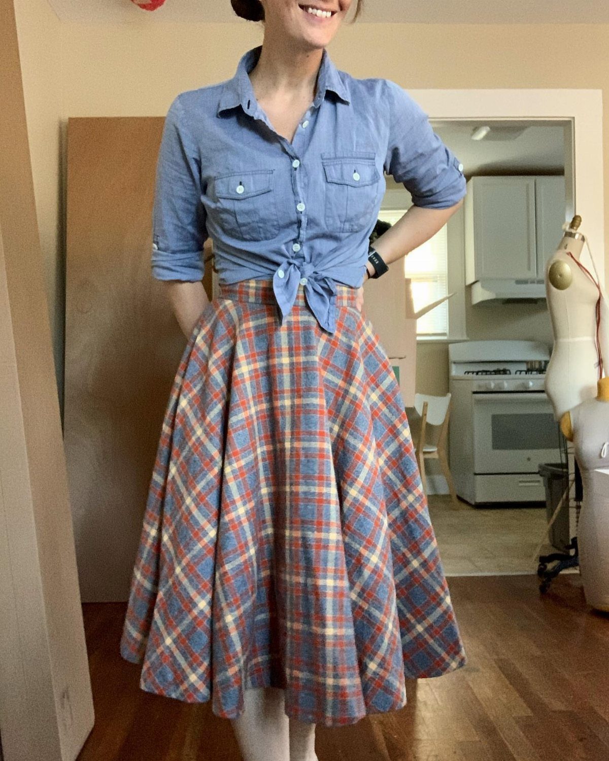 A woman wearing a blue denim shirt and a plaid flared midi skirt standing indoors.