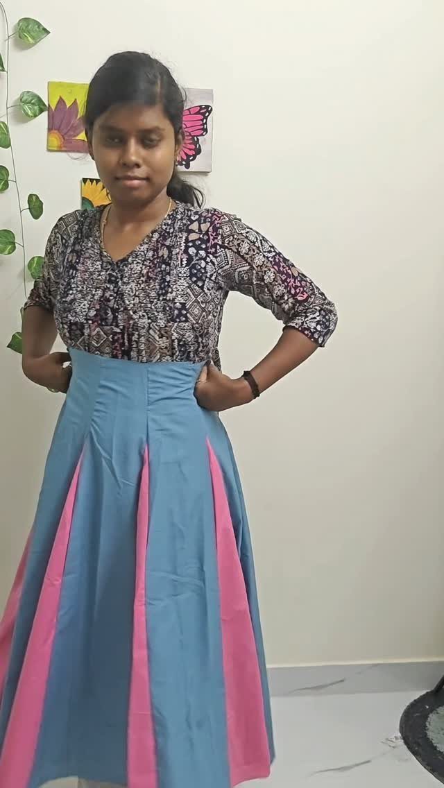 A woman posing in a long blue and pink paneled skirt against a white wall.