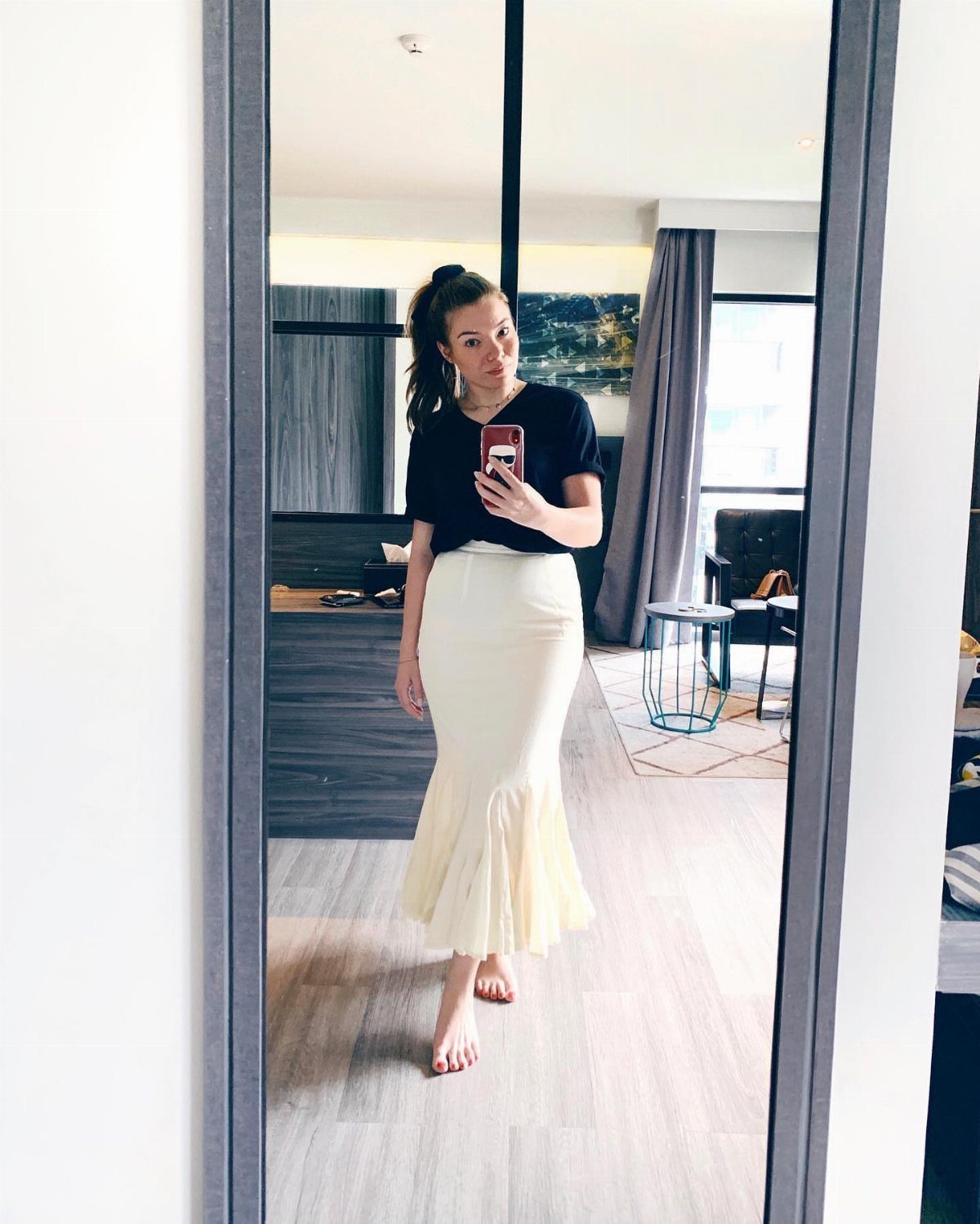 A woman posing in a mirror wearing a cream-colored mermaid style skirt and black t-shirt.