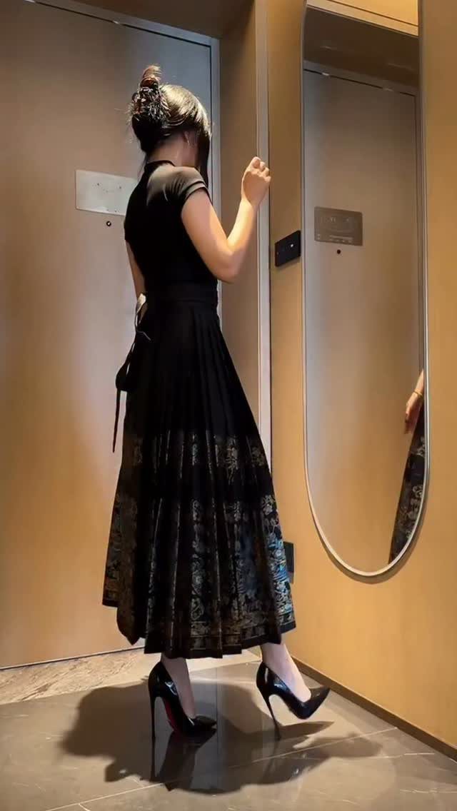 Woman in a black pleated midi skirt and high heels standing in a hallway.