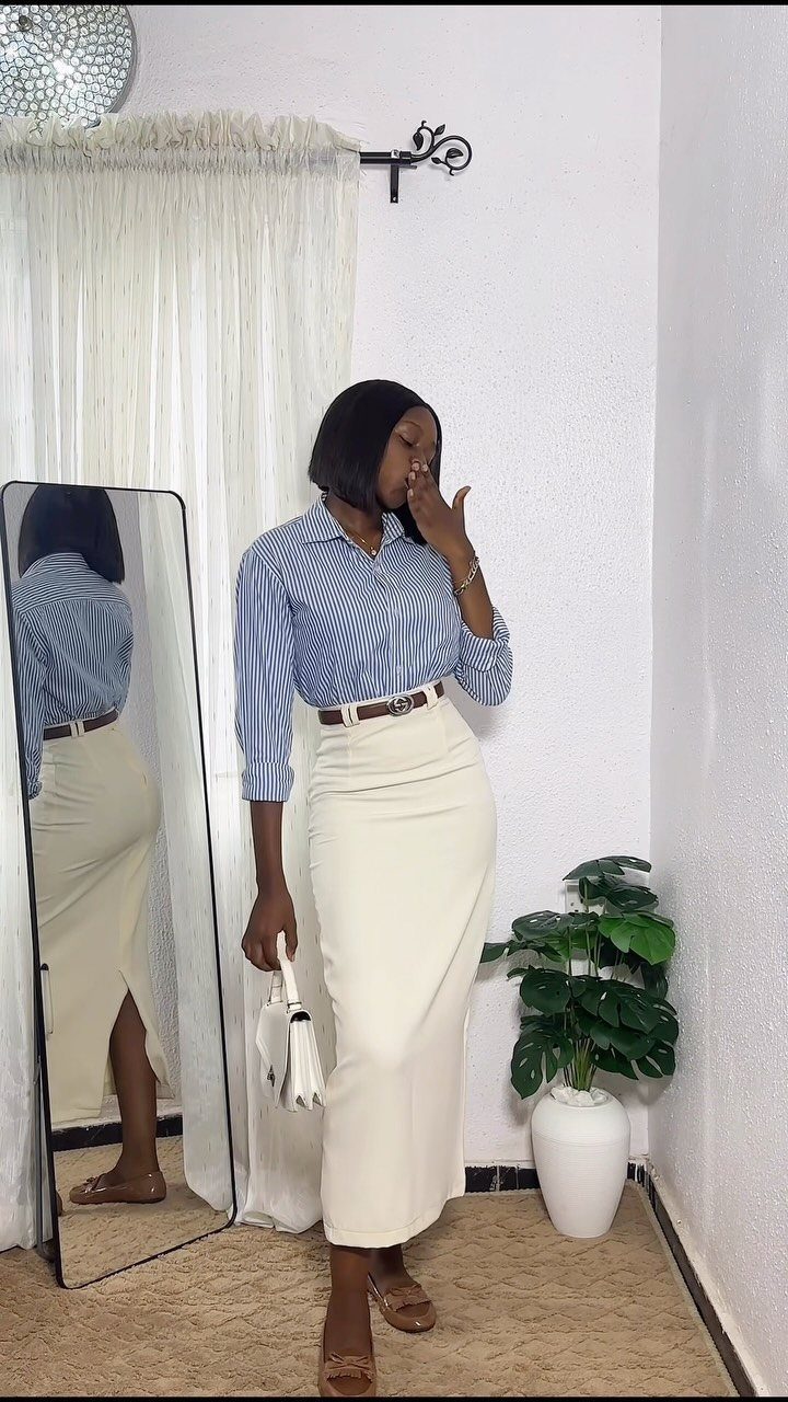 Woman wearing a long cream-colored straight skirt and a blue checkered shirt standing near a mirror.