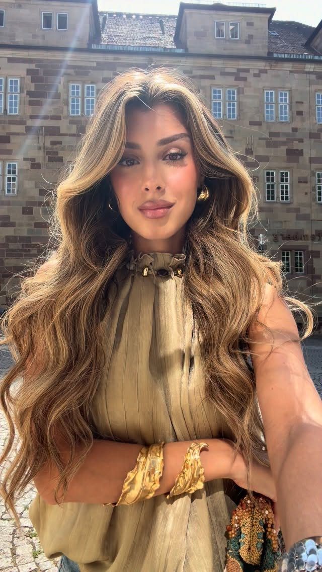 A woman with long wavy hair wearing chunky gold bracelets and earrings outdoors.