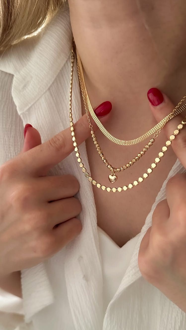 Three layered gold necklaces with a heart pendant worn over a white linen shirt.
