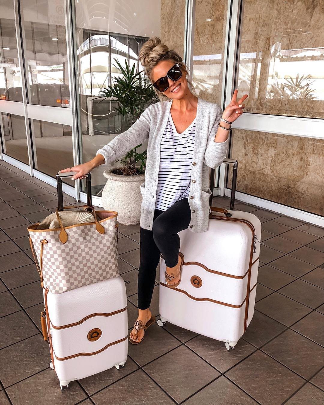 A woman in a grey cardigan and striped top poses with white suitcases.