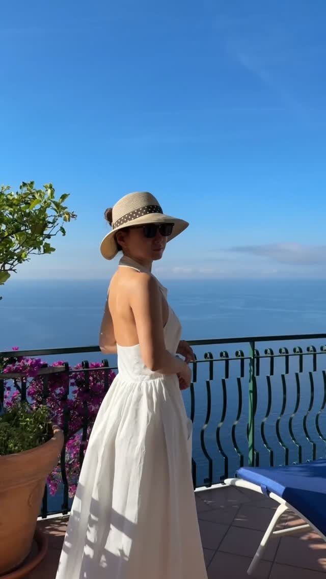 A woman in a white dress wearing a straw sun hat on a balcony overlooking the sea.