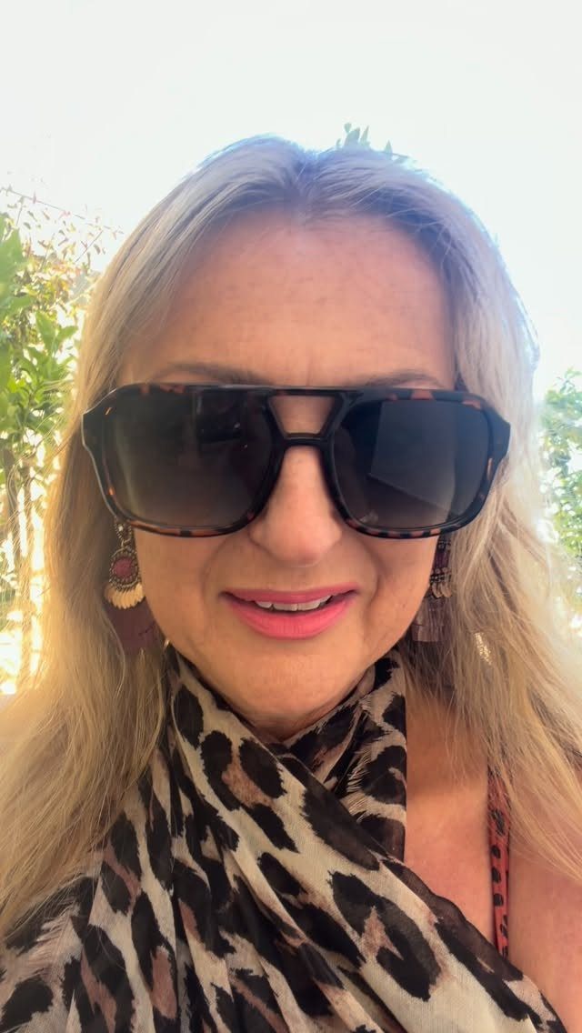 A blonde woman wearing large tortoiseshell sunglasses and a leopard print top outdoors.